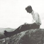 Benton MacKaye in a black-and-white photograph sitting atop a rocky overlook, looking down at a notebook.