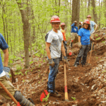 Group of 7 volunteers wearing hardhats and smiling at the camera, as they work on the Trail.